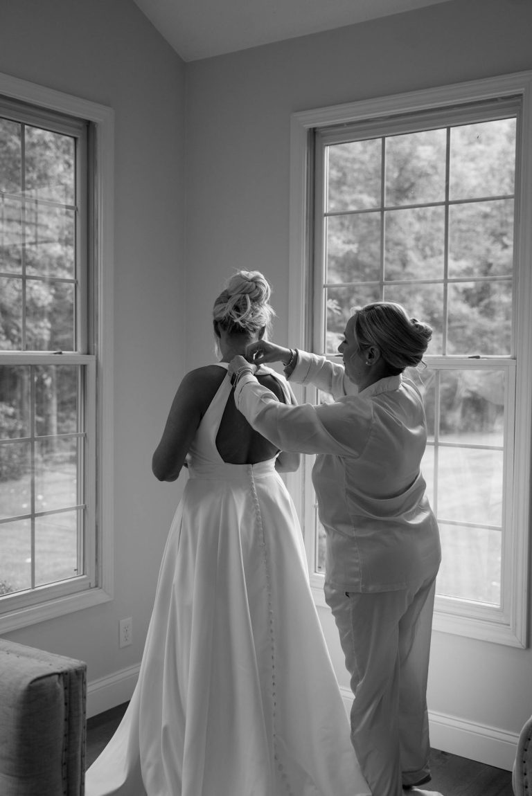 A bride prepares for her wedding as her mother assists with her dress, creating an intimate moment.